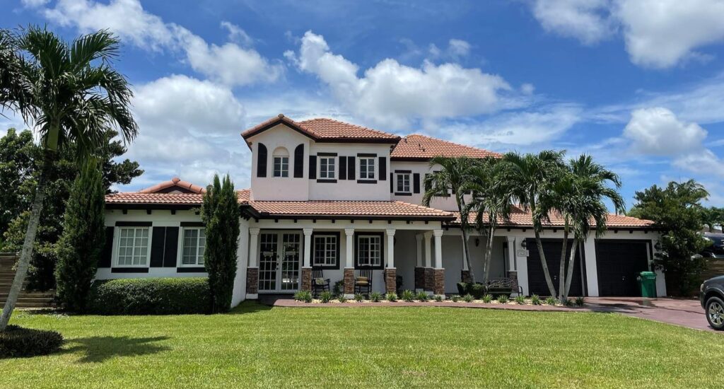 An exterior shot of a large, Mediterranean-style house with a clean terracotta roof, white walls, and dark shutters under a bright blue sky.