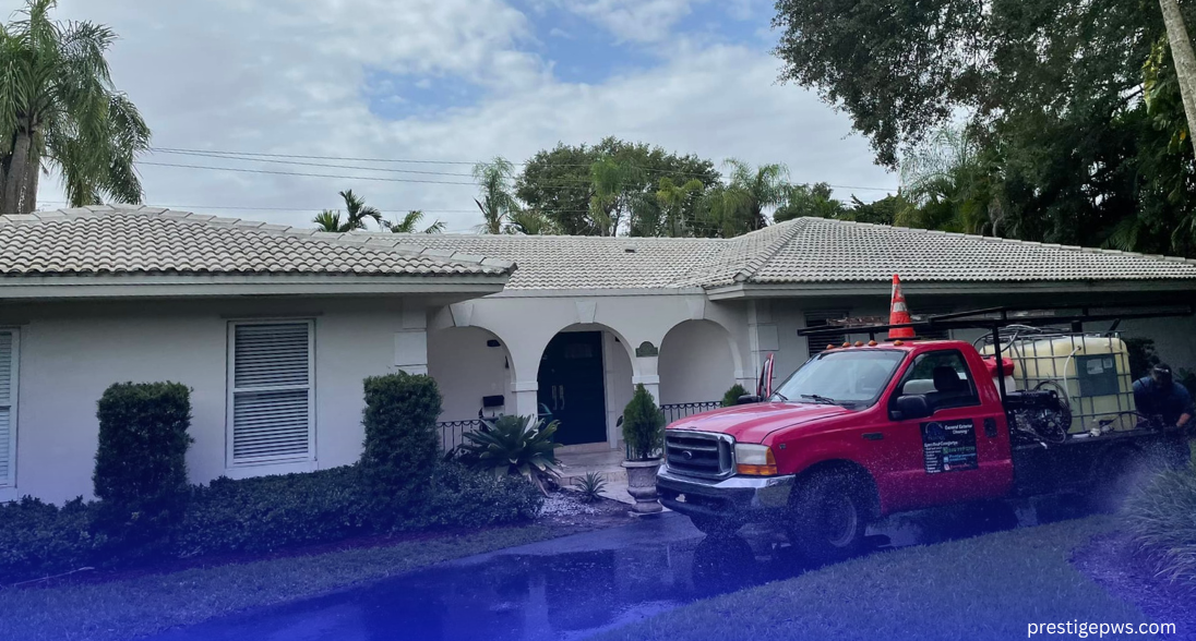 A red flatbed truck with cleaning equipment is parked on a wet driveway in front of a white, ranch-style house with a tile roof and arched entryways.