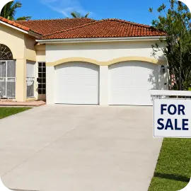 A freshly cleaned concrete driveway leading to a white two-car garage with a "For Sale" sign on the front lawn.