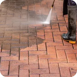 A close-up of a worker in rubber boots power washing a brick paver driveway, blasting away dark dirt and grime to reveal the clean surface beneath.