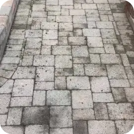 A stone paver walkway with dark embedded stains, dirt buildup, and weathered patches between the blocks.