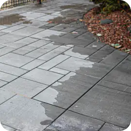 Dark gray interlocking stone pavers showing a distinct contrast between damp areas and dull, faded dry surfaces.
