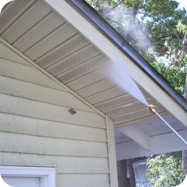 A high-pressure water spray cleaning the cream-colored siding and soffit of a house.