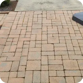 An unsealed interlocking stone paver patio showing faded color and small weeds growing within the joint lines.