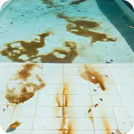 A tiled pool deck and steps heavily discolored with deep brown chemical stains and dark residues next to the water.