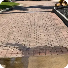 A weathered brick paver driveway and patio area showing surface discoloration, fading, and damp stains near the curb.